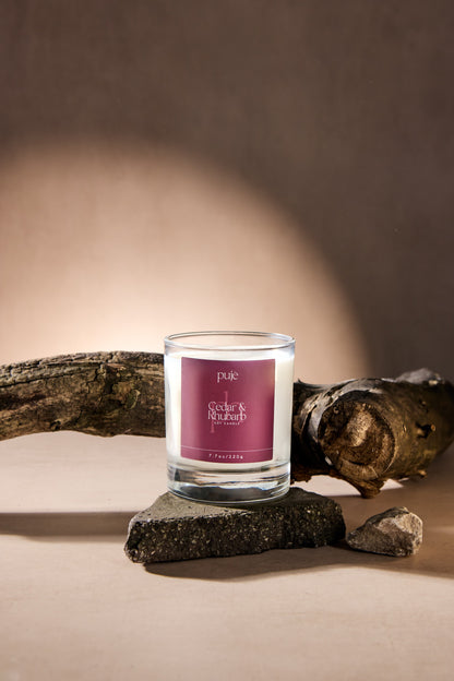 A glass soy wax candle with a burgundy label that reads "Cedar & Rhubarb Scented.